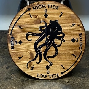 May include: A wooden clock with a black octopus design. The clock face has markings for high tide and low tide, with numbers 0-5 on each side. The text "HIGH TIDE" and "LOW TIDE" are printed on the clock face.
