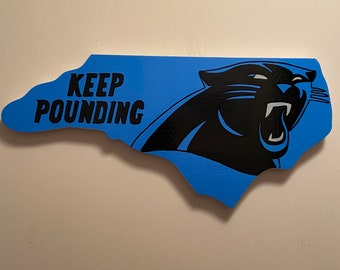 May include: A blue and black wooden sign shaped like the state of North Carolina with the words "Keep Pounding" and a black panther head.