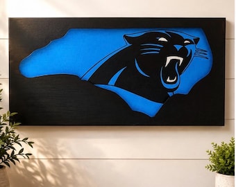 Könnte beinhalten: Ein rechteckiges schwarzes Wandschild mit einem Ausschnitt in Form des Bundesstaates North Carolina. Der Ausschnitt zeigt einen leuchtend blauen Hintergrund mit einem schwarz-weißen Pantherkopf-Logo, das in der Silhouette zentriert ist.