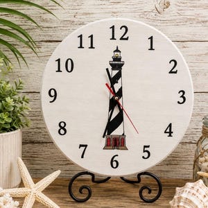 May include: A round white analog clock featuring black numbers from 1 to 12 and a central illustration of a black and white striped lighthouse with a red base. The clock has black hour and minute hands and a thin red second hand on a black stand.
