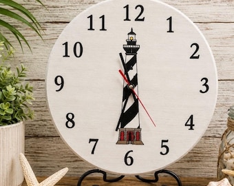 May include: A round white analog clock featuring black numbers from 1 to 12 and a central illustration of a black and white striped lighthouse with a red base. The clock has black hour and minute hands and a thin red second hand on a black stand.