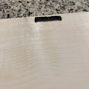 May include: Close-up of the back of a light-colored wooden sign with a black sawtooth hanger attached at the top. The wood grain is visible, and the surface has a slightly textured appearance.