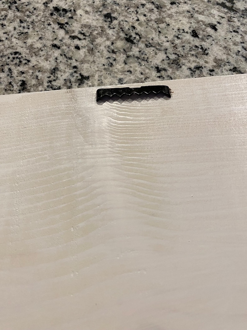 May include: Close-up of the back of a light-colored wooden sign with a black sawtooth hanger attached at the top. The wood grain is visible, and the surface has a slightly textured appearance.