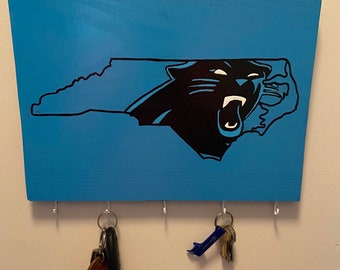May include: A blue wooden key holder with a black silhouette of a panther in the shape of North Carolina. The key holder has five hooks for hanging keys.