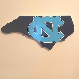 Pu&ograve; includere: Un contorno in legno dello stato della Carolina del Nord dipinto in blu scuro con un logo "NC" blu chiaro e bianco al centro.