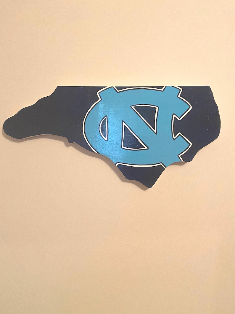 Pu&ograve; includere: Un contorno in legno dello stato della Carolina del Nord dipinto in blu scuro con un logo "NC" blu chiaro e bianco al centro.