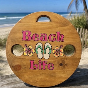 May include: A round wooden wine table with a handle cutout and two circular glass holders. The surface features the pink text Beach Life, a pair of cream flip-flops with green straps, and two yellow and orange hibiscus flowers printed on it.