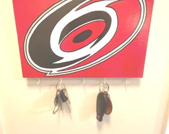 May include: A red wooden key holder with a black and white Carolina Hurricanes logo. The key holder has five hooks for hanging keys.