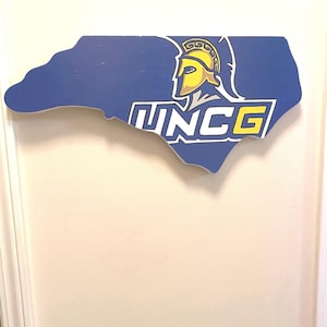 May include: A blue wooden cutout of the state of North Carolina with the UNCG logo. The logo features a Spartan helmet in yellow and a blue and white text that reads "UNCG".