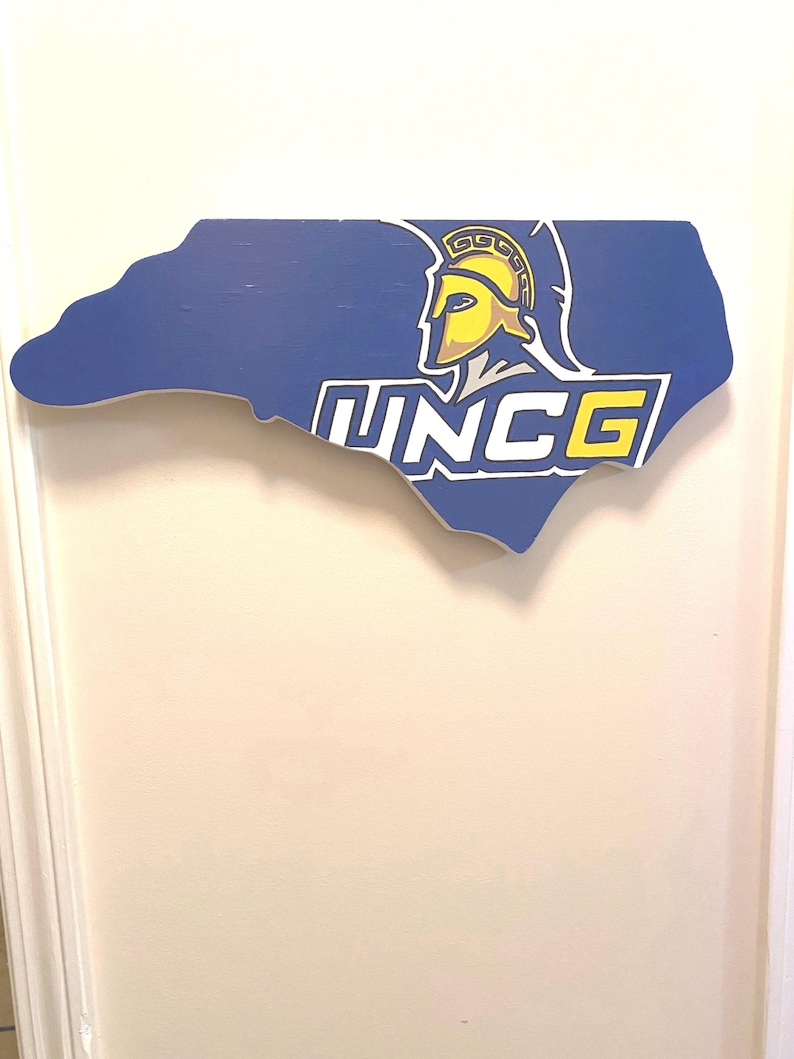 May include: A blue wooden cutout of the state of North Carolina with the UNCG logo. The logo features a Spartan helmet in yellow and a blue and white text that reads "UNCG".