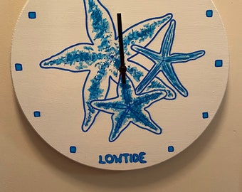 May include: A white round clock with blue starfish painted on it. The clock face reads "HIGH TIDE" at the top and "LOW TIDE" at the bottom.