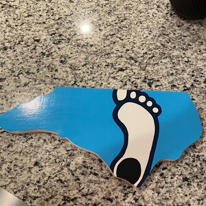 May include: A blue and white wooden cutout of the state of North Carolina with a white footprint in the shape of the state. The footprint has a black heel.