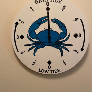 May include: A white circular tide clock with a blue crab illustration and black hands. The clock face reads "HIGH TIDE" and "LOW TIDE" with numbers 0 through 5.