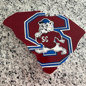May include: A red and blue wooden cutout of the state of South Carolina with a cartoon bulldog wearing a blue cap and red sweater with the letters "SC" on it.
