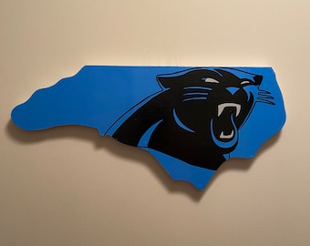 May include: A wooden sign in the shape of North Carolina, painted Carolina blue. The black silhouette of a panther with its mouth open is on the right side of the sign.