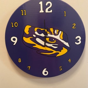 May include: A purple and yellow clock with the numbers 1 through 12 around the face. The clock has a black hand and a white background with a purple and yellow design.