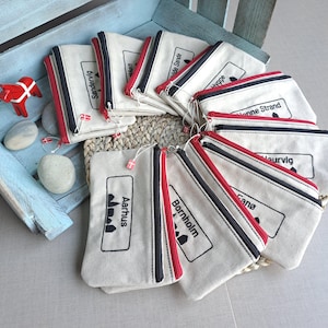 Könnte beinhalten: Eine Sammlung von beigefarbenen Canvas-Reißverschlusstaschen, die fächerförmig angeordnet sind. Jede Tasche hat ein schwarzes, besticktes rechteckiges Etikett mit einem Stadtnamen und einem kleinen Silhouetten-Symbol. Die Reißverschlüsse sind rot-schwarz.
