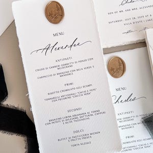 Może przedstawiać: Eleganckie karty menu weselnego z teksturowanego, złamanego białego papieru. Każda karta posiada złotą pieczęć woskową i ręcznie pisaną kaligrafię. Menu zawiera imię "Alexandra" i listę dań.