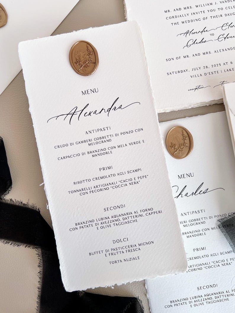 Może przedstawiać: Eleganckie karty menu weselnego z teksturowanego, złamanego białego papieru. Każda karta posiada złotą pieczęć woskową i ręcznie pisaną kaligrafię. Menu zawiera imię "Alexandra" i listę dań.