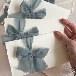 May include: Stack of white envelopes with frayed edges, each tied with a dusty blue ribbon bow.