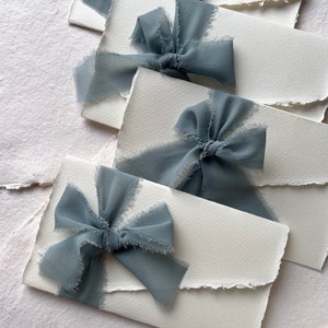 May include: Four white envelopes with frayed edges, each tied with a dusty blue ribbon bow.