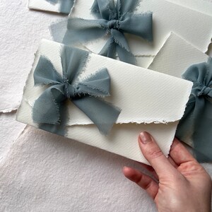 May include: White envelopes with frayed edges, decorated with dusty blue ribbon bows. The envelopes are stacked on top of each other.