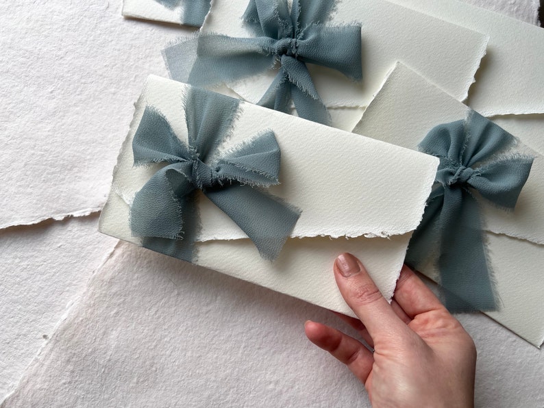 May include: White envelopes with frayed edges, decorated with dusty blue ribbon bows. The envelopes are stacked on top of each other.