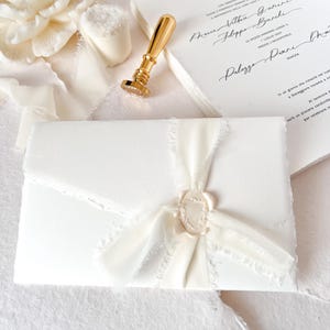 May include: A close-up of wedding stationery featuring a white envelope sealed with a cream-coloured wax seal and ribbon. A gold wax stamp and invitation cards with handwritten text are also visible. The overall theme is elegant and romantic.