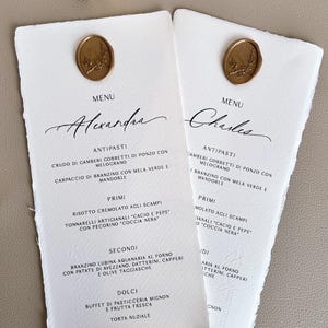 Może przedstawiać: Dwa białe papierowe menu z postrzępionymi krawędziami, z eleganckimi kaligraficznymi imionami "Alexandra" i "Charles". Każde menu jest ozdobione złotą pieczęcią woskową. Menu zawiera włoskie dania.