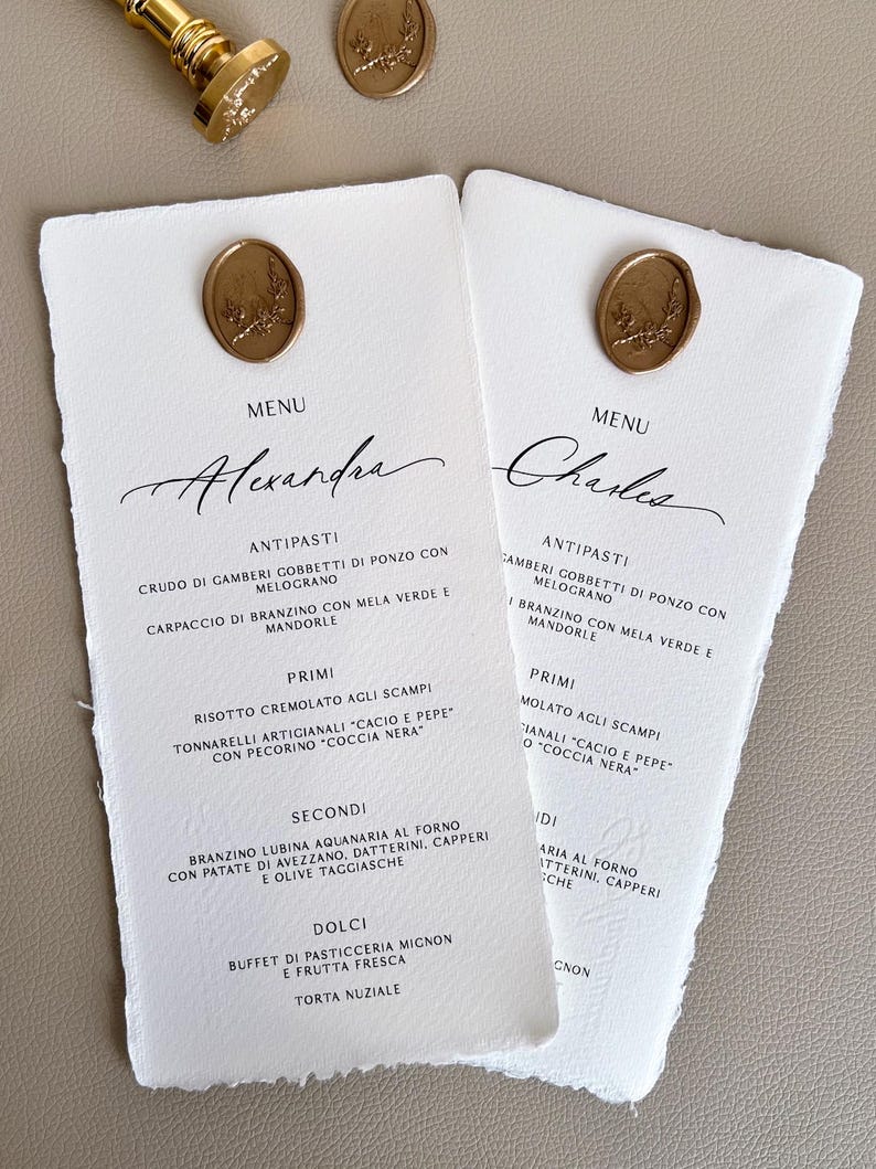 Może przedstawiać: Dwa białe papierowe menu z postrzępionymi krawędziami, z eleganckimi kaligraficznymi imionami "Alexandra" i "Charles". Każde menu jest ozdobione złotą pieczęcią woskową. Menu zawiera włoskie dania.