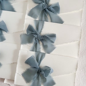 May include: Stack of white envelopes with a dusty blue ribbon tied in a bow on each envelope.
