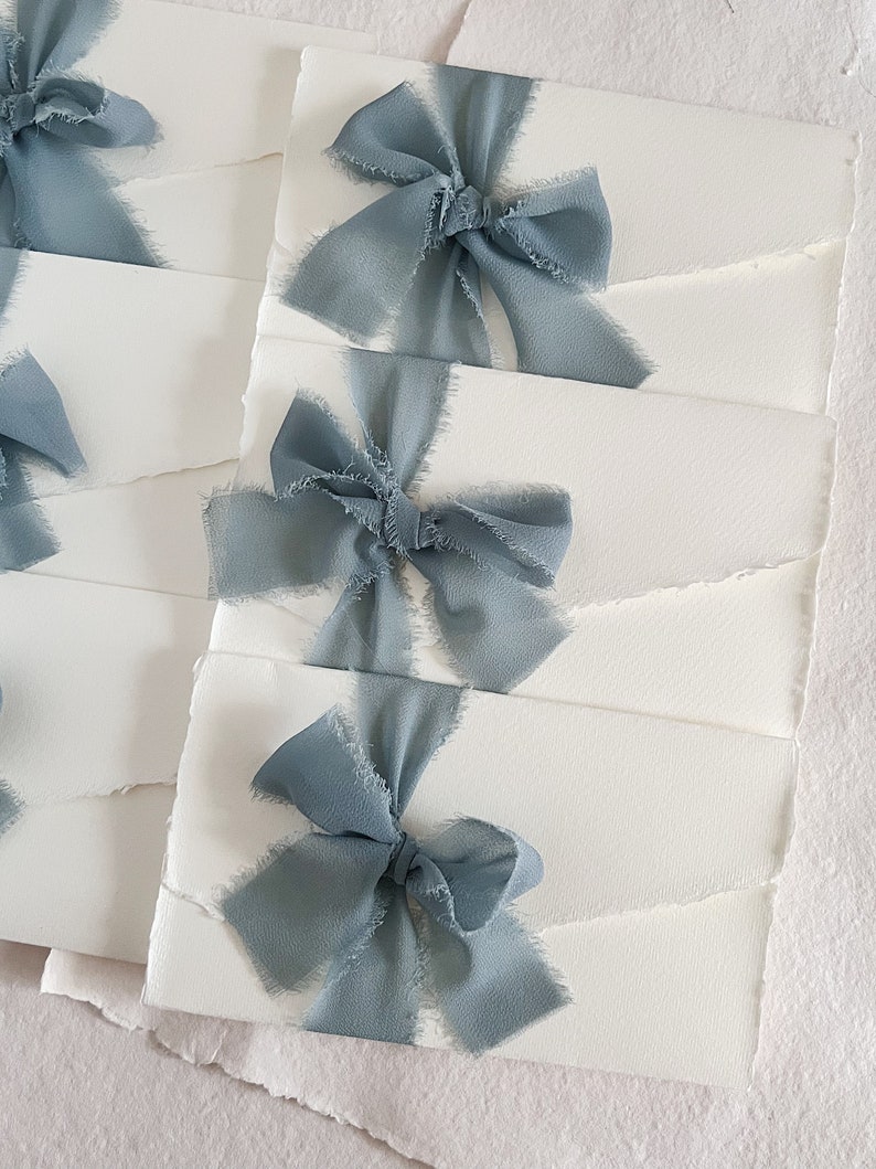 May include: Stack of white envelopes with a dusty blue ribbon tied in a bow on each envelope.