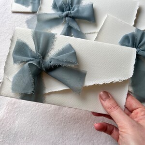 May include: A white envelope with a frayed edge, tied with a light blue ribbon bow. The envelope is on a white background.