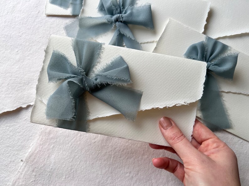 May include: A white envelope with a frayed edge, tied with a light blue ribbon bow. The envelope is on a white background.