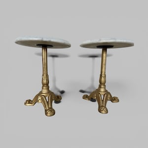 May include: A pair of small side tables, each featuring a circular white marble top supported by a single central pedestal column and a three-legged base finished in a metallic gold tone with ornate, textured detailing on the feet.