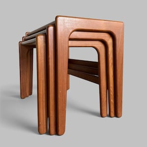 May include: A set of three nested tables crafted from medium-toned wood with a visible grain. Each table features a rectangular top and four legs with rounded edges, designed to slide neatly underneath one another for compact storage.