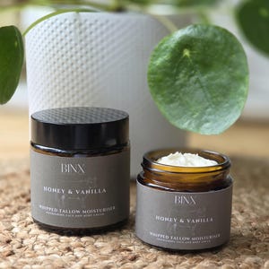 Op de afbeelding: Twee amberkleurige glazen potten met Binx Honey & Vanilla Whipped Tallow Moisturiser. Eén pot is afgesloten met een zwarte deksel, de andere is open en toont witte crème. De grijze etiketten bevatten witte tekst en een lijntekening van een koe.