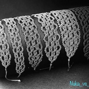 Op de afbeelding: Een set van zes zilverkleurige macramé armbanden met ingewikkelde knoop patronen. De armbanden worden getoond op een zwart cilindrisch oppervlak.