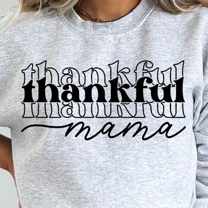Peut inclure: Un sweat-shirt gris clair avec l'inscription "thankful mama" en noir, utilisant des lettres script et en blocs. Le sweat-shirt a un col rond et des manches longues. Le bas de l'image affiche "SVG. JPEG. PNG. EPS. DXF."