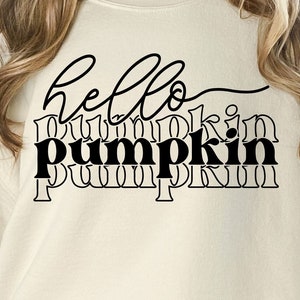 Peut inclure: Un sweat-shirt crème avec les mots "hello pumpkin" en écriture noire et en lettres bâtons. Le sweat-shirt est présenté devant une fenêtre avec vue sur des arbres. En dessous du sweat-shirt, on peut lire "SVG. JPEG. PNG. EPS. DXF."