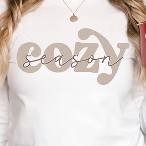Puede incluir: Camiseta blanca de manga larga con las palabras "cozy season" en color topo y fuente manuscrita. Se sostiene una taza roja con "warm cozy" en escritura blanca. Un árbol de Navidad con adornos está en el fondo.
