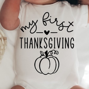 Pode incluir: Macacão branco para bebê com texto preto que diz "my first thanksgiving" com um pequeno coração entre as palavras. Um contorno preto de uma abóbora está abaixo do texto.