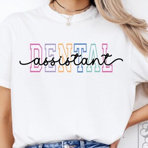 May include: White t-shirt with the words "DENTAL assistant" in colorful block letters and a cursive font. The shirt is a casual style, suitable for everyday wear. The design is centered on the chest.