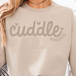 Puede incluir: Sudadera beige con la frase "cuddle season" en una fuente suave y cursiva. La palabra "cuddle" tiene pequeños acentos de corazón. La sudadera es de color neutro y tiene un ajuste relajado.