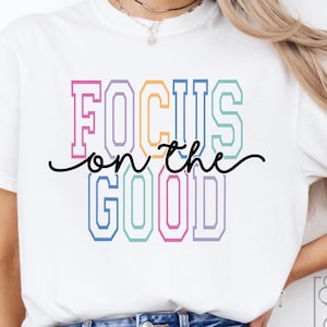 Puede incluir: Camiseta blanca con la frase "FOCUS on the GOOD" en letras de bloque coloridas y una fuente cursiva. La camiseta tiene cuello redondo y mangas cortas. El diseño utiliza tonos de rosa, naranja, verde y azul.