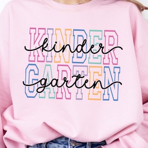 May include: A light pink sweatshirt with the words "Kindergarten" in colorful block letters. The word "kinder" is above "garten" in a cursive font. The sweatshirt is a casual style, suitable for everyday wear.