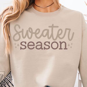 Puede incluir: Sudadera beige con las palabras "Sweater season" en una escritura marrón claro en la parte delantera. La palabra "season" está en una fuente marrón más oscura. Pequeñas explosiones de estrellas están a ambos lados de la palabra "season".