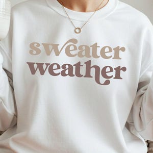 Puede incluir: Sudadera blanca con las palabras "sweater weather" en letras marrones y color topo. Una persona sostiene una taza beige en su mano izquierda y hace un signo de la paz con su mano derecha. La sudadera tiene mangas largas.