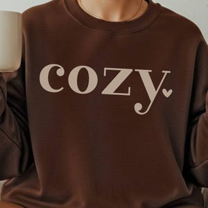 Puede incluir: Una sudadera marrón con la palabra "cozy" impresa en color beige claro. La palabra tiene un símbolo de corazón al final. Una persona sostiene una taza blanca en su mano izquierda y hace un signo de paz con su mano derecha.