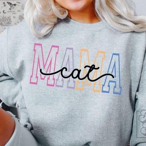 Könnte beinhalten: Ein hellgraues Sweatshirt mit den Worten "MAMA cat" in bunten Blockbuchstaben. Das Wort "cat" ist in einer Schreibschrift geschrieben. Das Sweatshirt ist ein lässiger Stil.